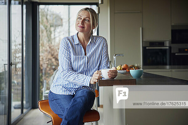 Thoughtful blond woman with coffee cup sitting in kitchen at home
