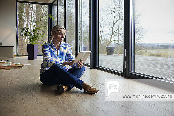 Woman using tablet PC sitting on floor by glass window at home