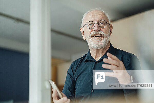 Smiling senior businessman holding smart phone gesturing in office