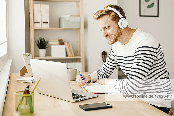 Smiling man wearing wireless headphones looking at laptop in living room