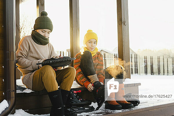 Junge und Bruder tragen Schlittschuhe und sitzen auf der Veranda eines Hauses im verschneiten Garten