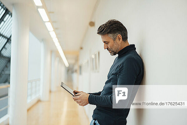 Businessman using tablet computer leaning on wall in corridor