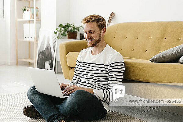 Smiling young man using laptop sitting cross-legged in front of sofa at home