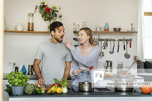 Smiling young woman feeding food to boyfriend in kitchen at home