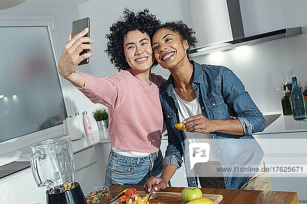 Happy friends taking selfie though smart phone standing in kitchen at home