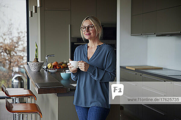 Woman holding coffee cup standing in kitchen at home