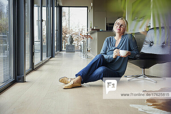 Thoughtful blond woman holding coffee cup sitting by chair at home
