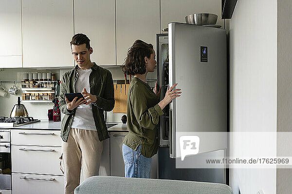 Junger Mann benutzt Tablet-PC  während Frau zu Hause den Kühlschrank in der Küche öffnet