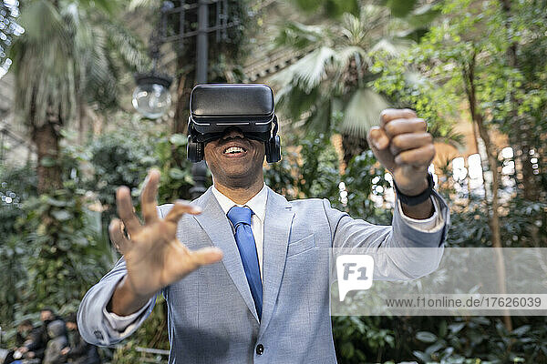 Smiling businessman wearing virtual reality simulator gesturing in front of plants