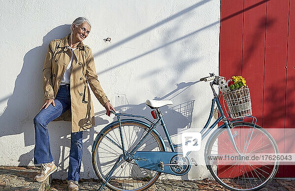 Smiling woman with bicycle standing in front of wall on sunny day