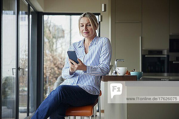 Blonde Frau surft mit Smartphone im Internet und sitzt in der Küche