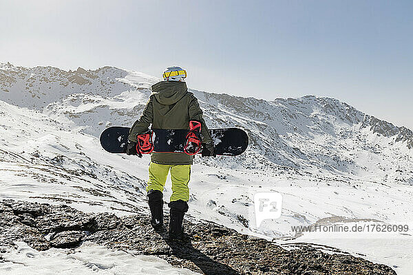 Junger Mann steht mit Snowboard auf einem Felsen