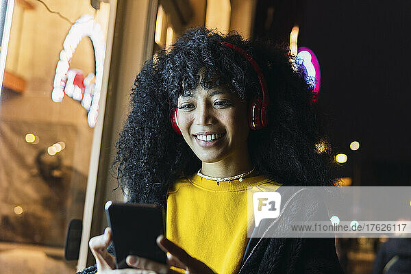 Happy young woman wearing wireless headphones using mobile phone at night
