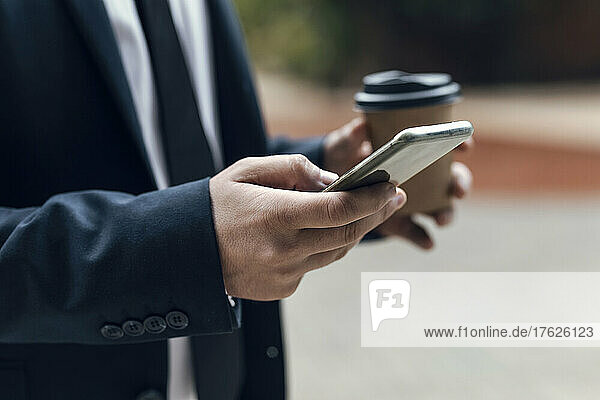 Hands of businessman using smart phone while holding disposable coffee cup