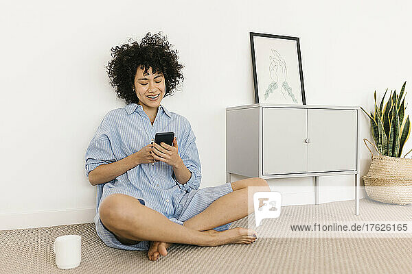 Happy young woman using smart phone sitting cross-legged at home