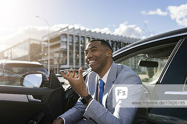 Cheerful businessman sending voicemail through mobile phone sitting in car on sunny day