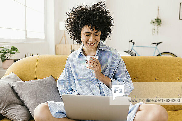 Smiling young woman with coffee cup using laptop at home