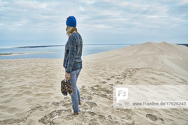 Woman holding sandals standing on sand dune at beach