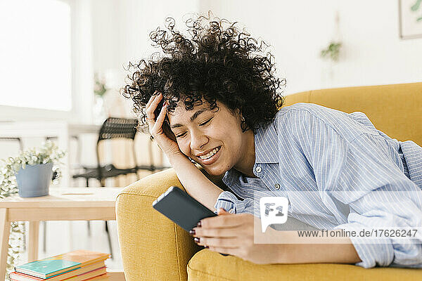 Happy woman with hand in hair using mobile phone on sofa at home