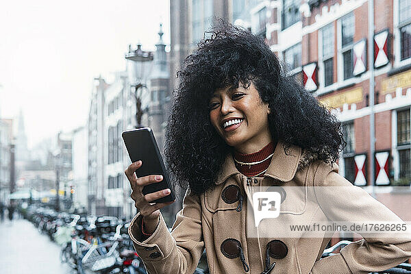 Cheerful young woman with tousled hair using smart phone in city