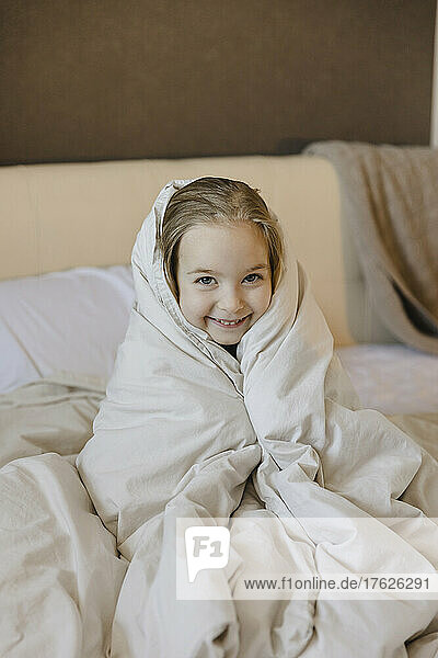 Smiling cute girl wrapped in blanket sitting on bed at home