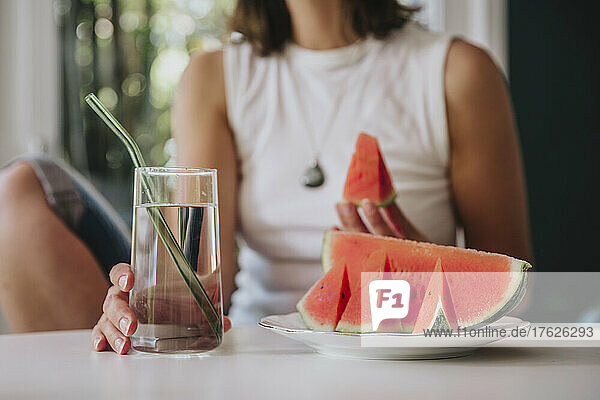 Woman holding drinking glass by watermelon slice in plate on table at home