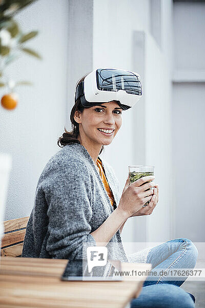 Lächelnde Geschäftsfrau im Virtual-Reality-Simulator mit Trinkglas auf Stuhl sitzend
