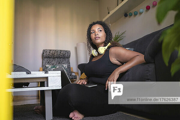 Young woman wearing wireless headphones sitting with laptop by sofa at home