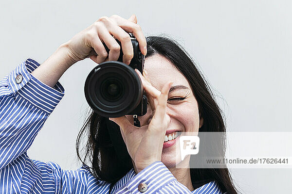Lächelnde Frau fotografiert mit der Kamera  die vor einer weißen Wand steht