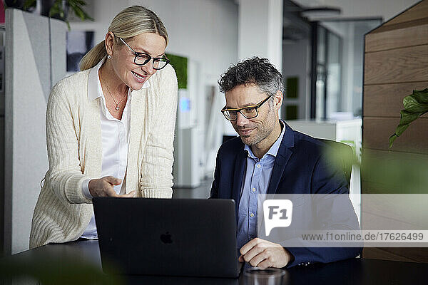 Lächelnde Geschäftsfrau diskutiert mit Kollegin über Laptop am Schreibtisch im Büro