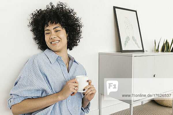 Happy young woman with coffee cup sitting by white wall at home