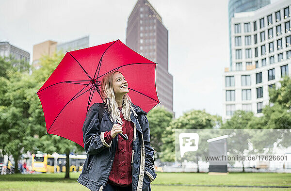 Glückliche junge Frau mit rotem Regenschirm steht im öffentlichen Park