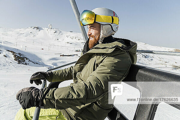 Smiling young man wearing jacket with ski goggles and helmet sitting on ski lift