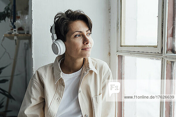 Contemplative woman with headphones listening to music by window