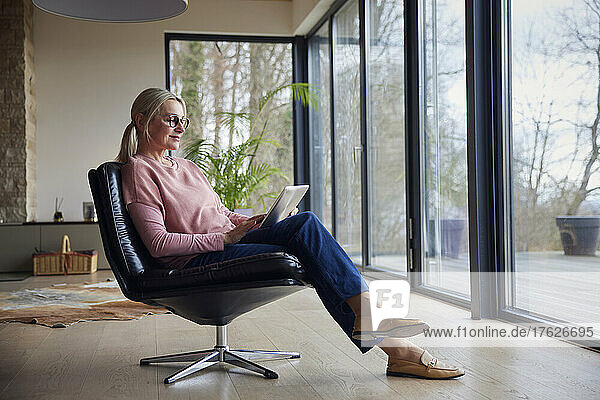 Smiling woman using laptop sitting on chair by glass window at home