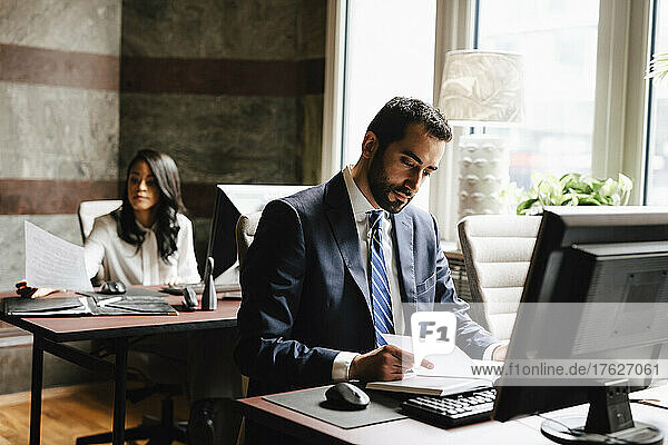 Selbstbewusster männlicher Anwalt liest Tagebuch mit einer weiblichen Kollegin  die im Hintergrund im Büro arbeitet