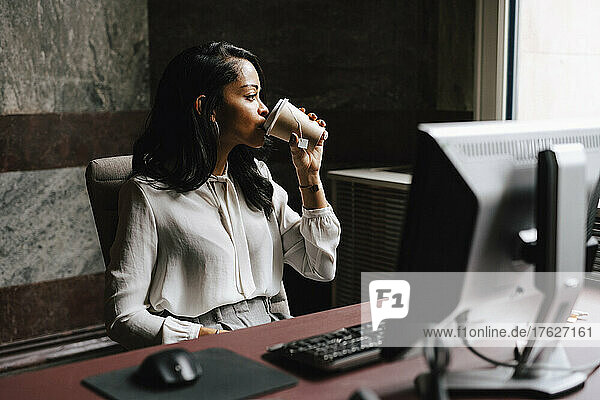 Female lawyer drinking coffee from disposable cup while sitting at desk in office