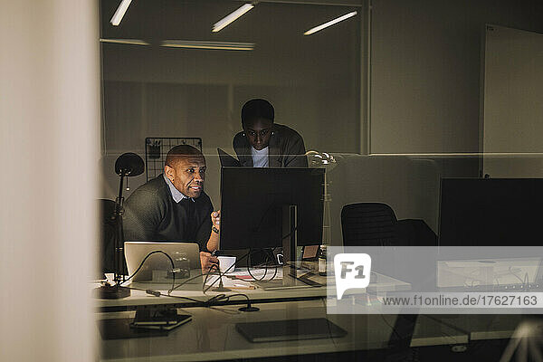 Businessman discussing with female colleague over computer seen through glass at work place