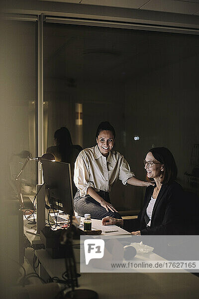 Smiling female colleagues discussing on computer while working last minute at work place
