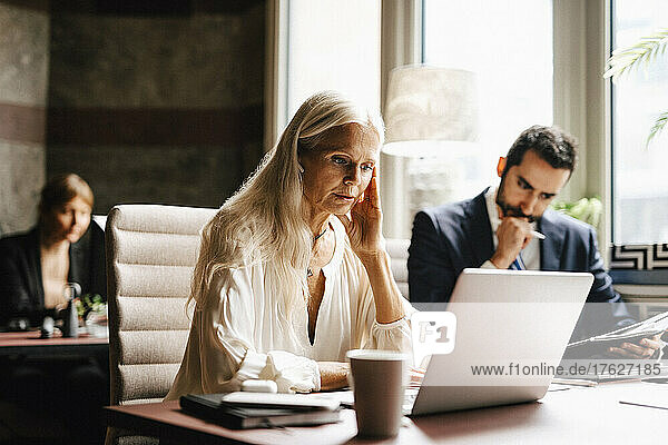 Mature businesswoman wearing in-ear headphones looking at laptop by businessman in office