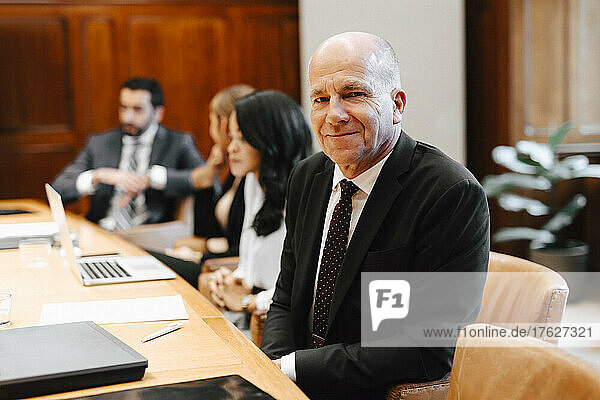 Portrait of smiling senior male lawyer in board room with colleagues during meeting