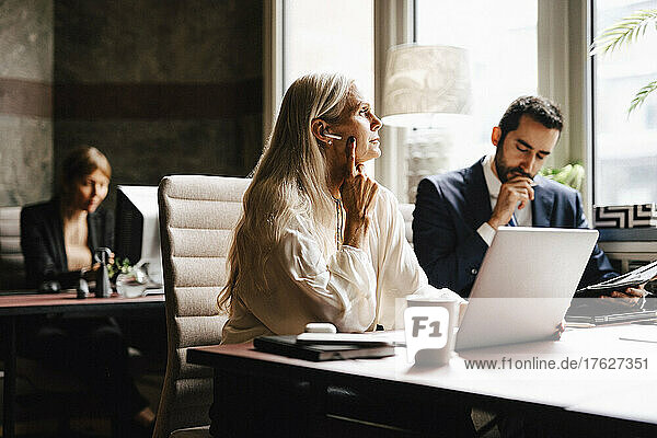 Reife Geschäftsfrau mit In-Ear-Kopfhörern am Laptop von Geschäftsmann am Schreibtisch im Büro