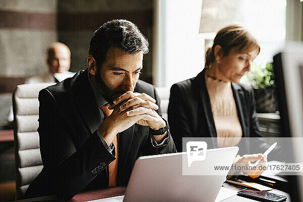 Businessman with hands clasped looking at laptop working by businesswoman at desk in law office