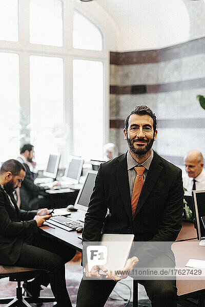 Portrait of smiling male lawyer holding laptop sitting on desk in office