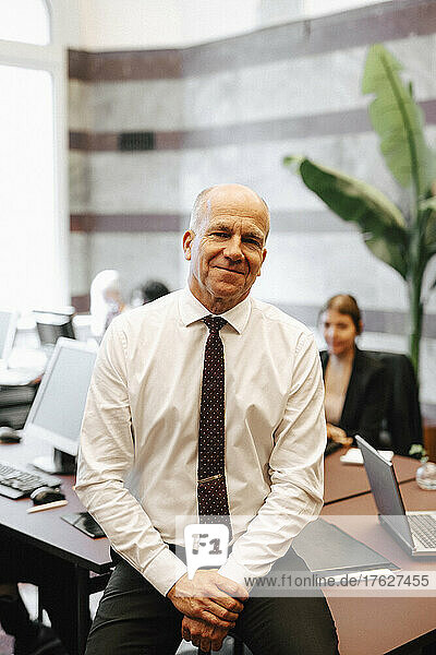 Porträt eines lächelnden älteren männlichen Anwalts  der in einem Büro am Schreibtisch sitzt