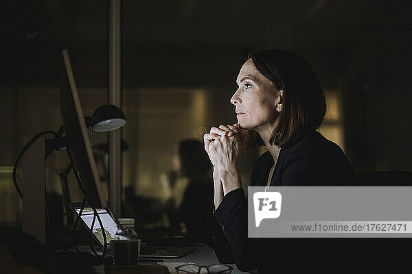 Mature businesswoman with hands clasped working over computer in office at night