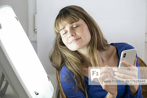 Woman sitting with her smartphone near a light therapy lamp.