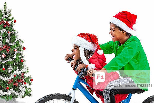 Smiling brother and sister wearing Santa's hat in front of Christmas tree on a blue bike decorated for Christmas.