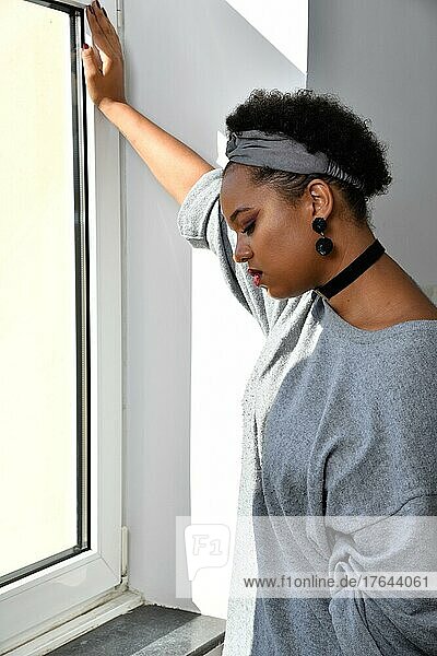 Eine nachdenkliche Afro-Amerikanische junge hübsche Frau steht an einem Fenster und blickt nachdenklich zu Boden. Sie trägt einen grauen Pullover mit einem passenden Stirnband  großem Ohrschmuck und einem schwarzen Halsband