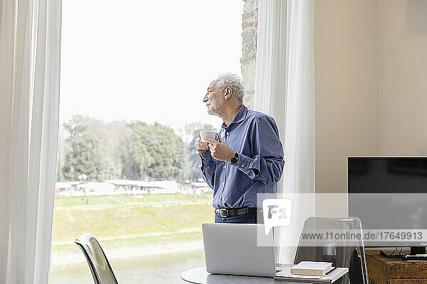 Älterer Mann mit Kaffeetasse  der durch das Fenster schaut und im Hotelapartment steht